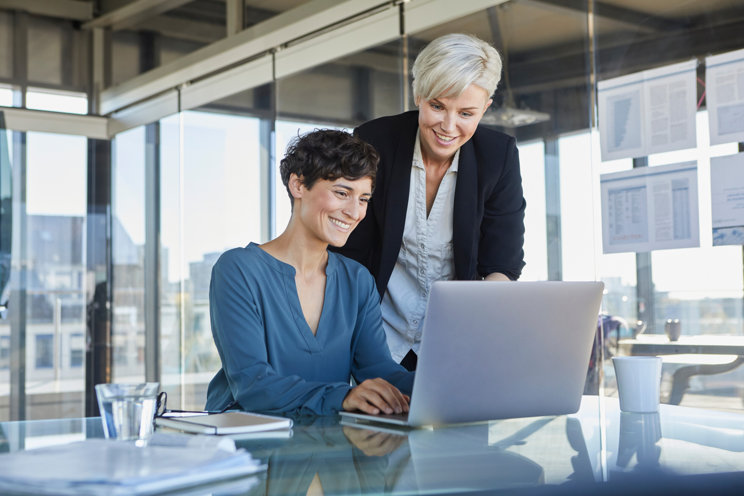 Two smiling businesswomen sharing laptop at desk in office